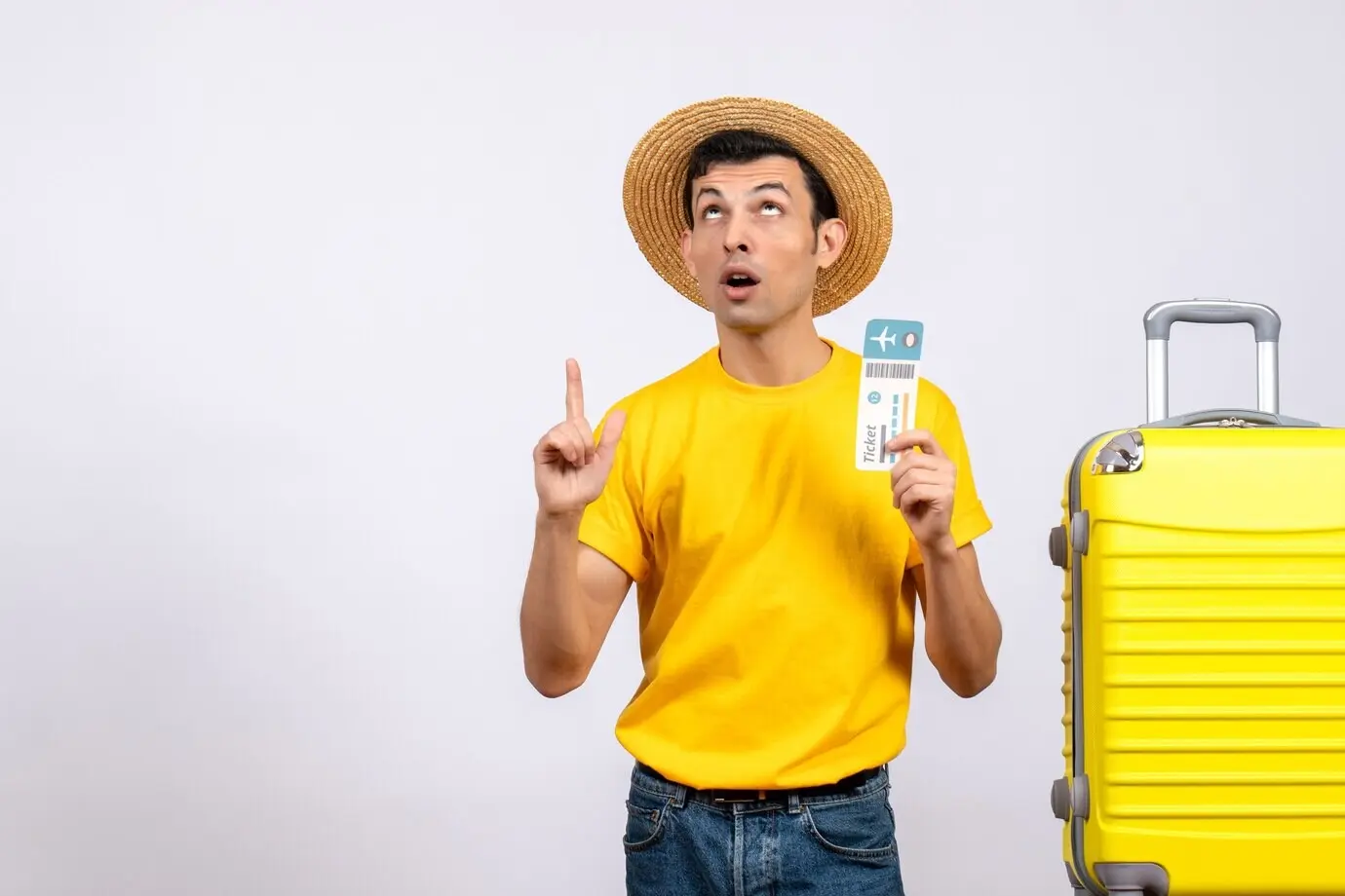 Front view of a young tourist in a yellow T-shirt standing next to a yellow suitcase, holding a ticket and pointing at the ceiling.