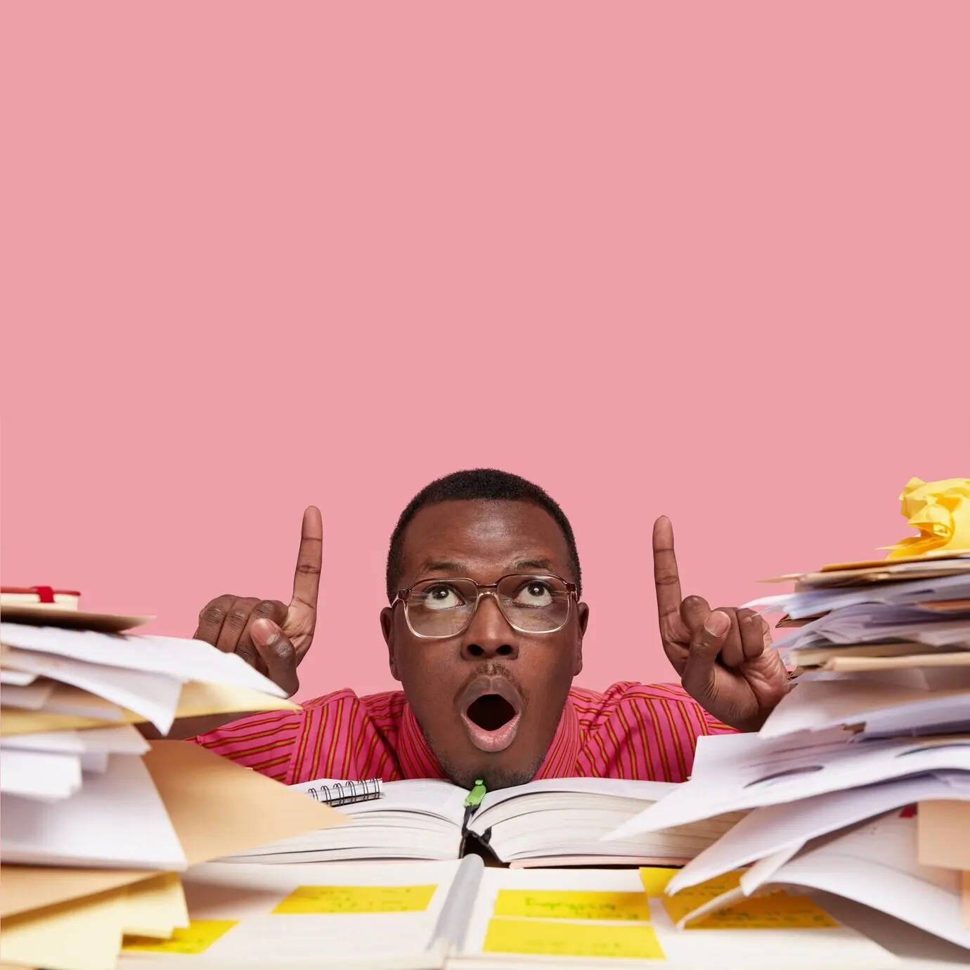 Vertical shot of a surprised black man with his mouth wide open, pointing both index fingers toward the ceiling, wearing glasses, with a lot of paperwork.